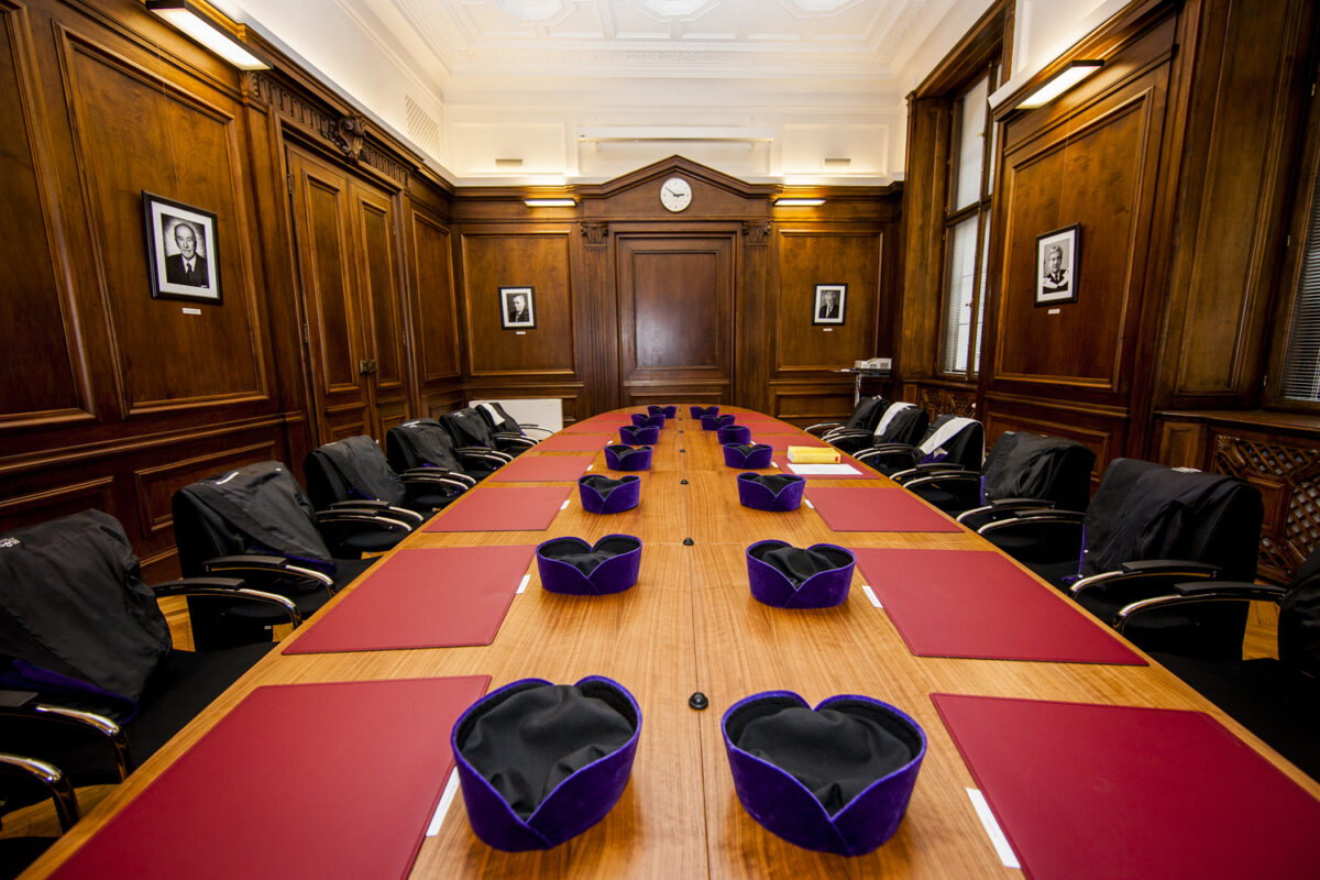 Ein Sitzungsraum des österreichischen Verfassungsgerichtshof. Eine lange Tafel mit Stühlen, roten Schreibunterlagen und den aufgereihten Kopfbedeckungen der Richter*innen.