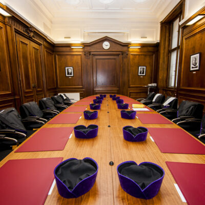 Ein Sitzungsraum des österreichischen Verfassungsgerichtshof. Eine lange Tafel mit Stühlen und mit roten Schreibunterlagen. Über den Rückenlehnen der Stühle hängen die Roben und auf dem Tisch liegen die aufgereihten Kopfbedeckungen der Richter*innen.