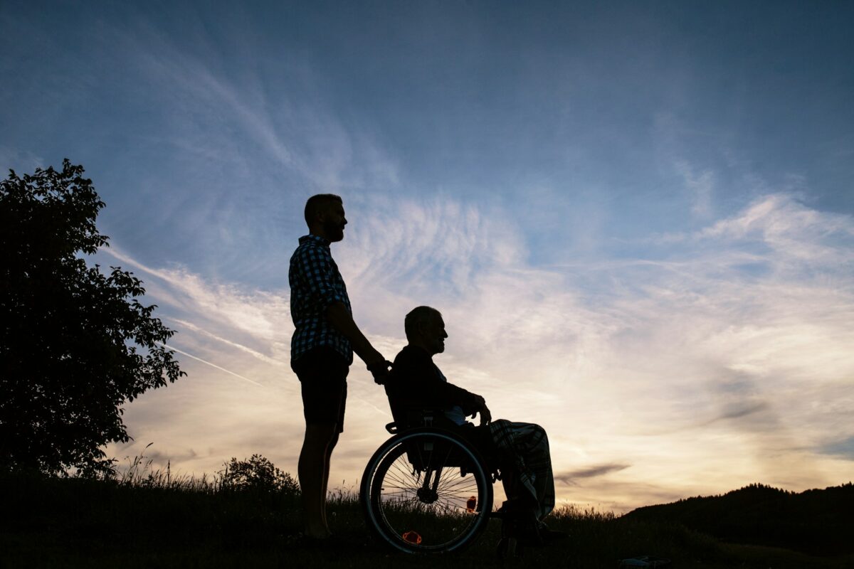 In der Abenddämmerung zeichnen sich die Silhouetten einer Person im Rollstuhl und einer schiebenden Person auf einer Wiese ab.