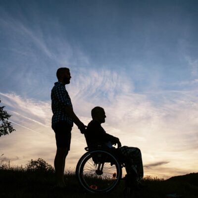 In der Abenddämmerung zeichnen sich die Silhouetten einer Person im Rollstuhl und einer schiebenden Person auf einer Wiese ab.